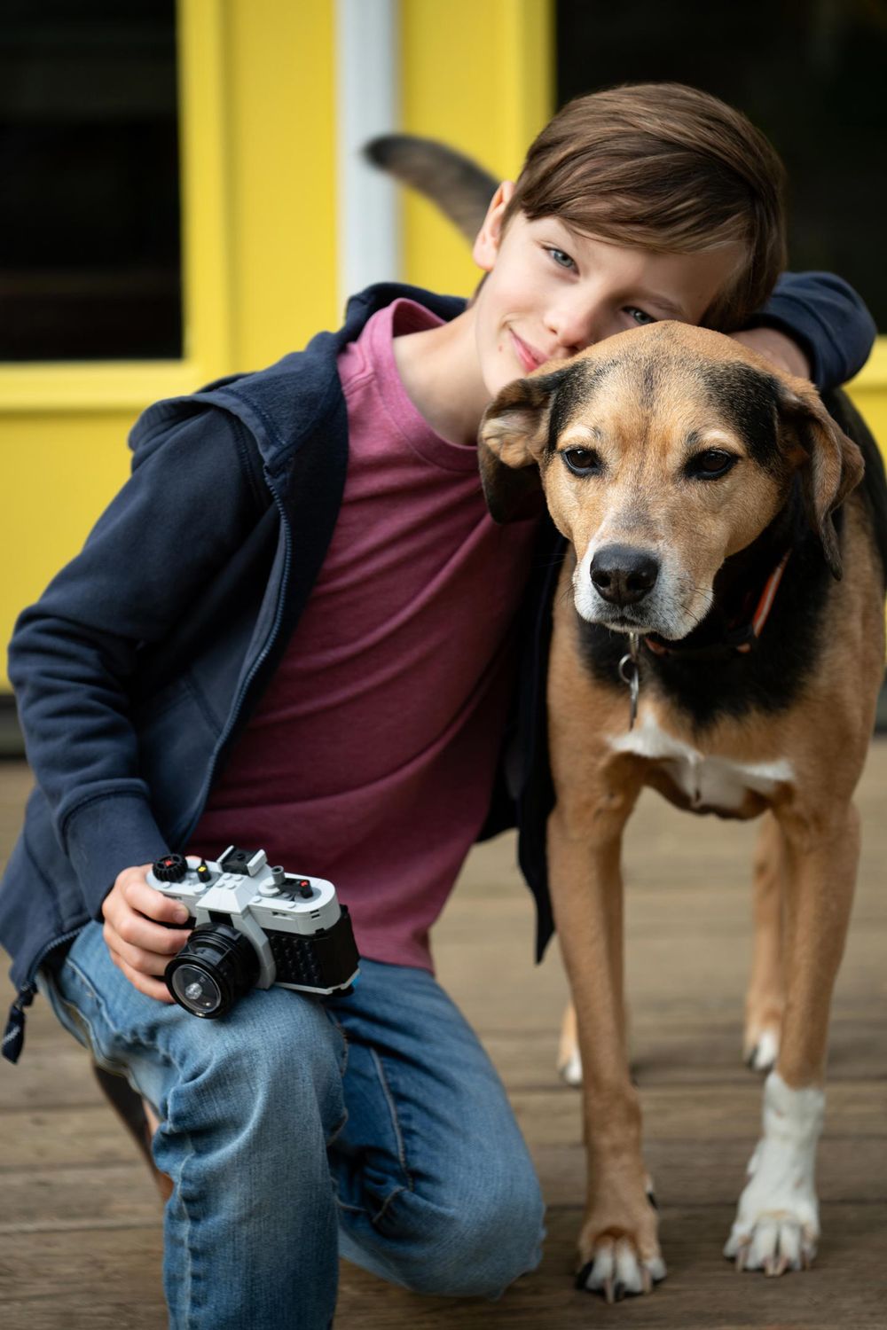 Young photographer with a vintage camera poses with a friendly brown and white mixed breed dog on wooden porch.