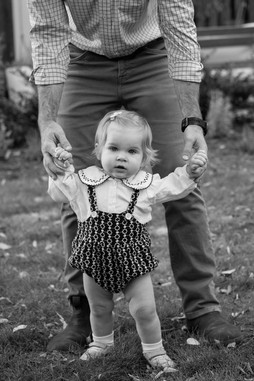 Black and white photo of small toddler in patterned romper learning to walk while holding adult's hands.