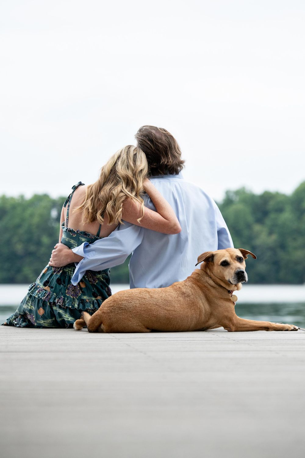 A couple sits together on a dock overlooking water with their brown dog resting beside them on a peaceful afternoon.
