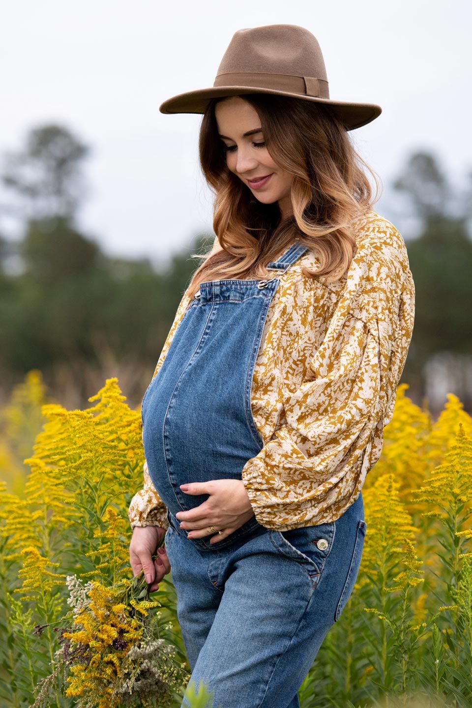 A maternity photo in a field of yellow flowers featuring denim overalls, brown hat and floral top.