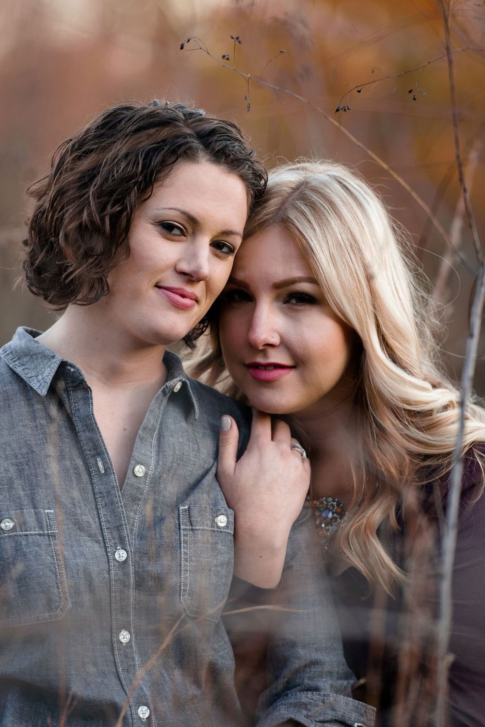 Two people pose together outdoors wearing matching denim shirts against an autumn background with soft bokeh lighting.