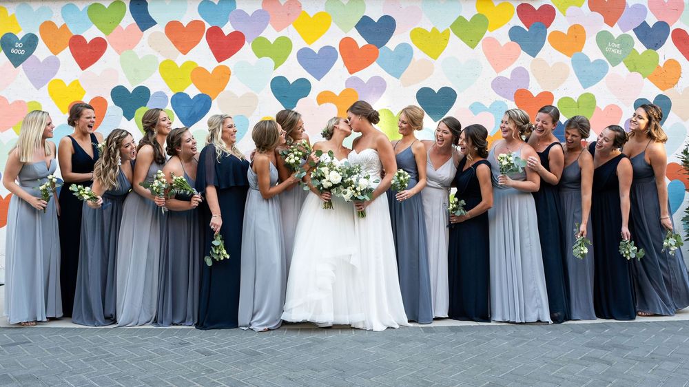 Wedding party members in coordinating blue and gray dresses stand together outdoors in a celebratory group photo.