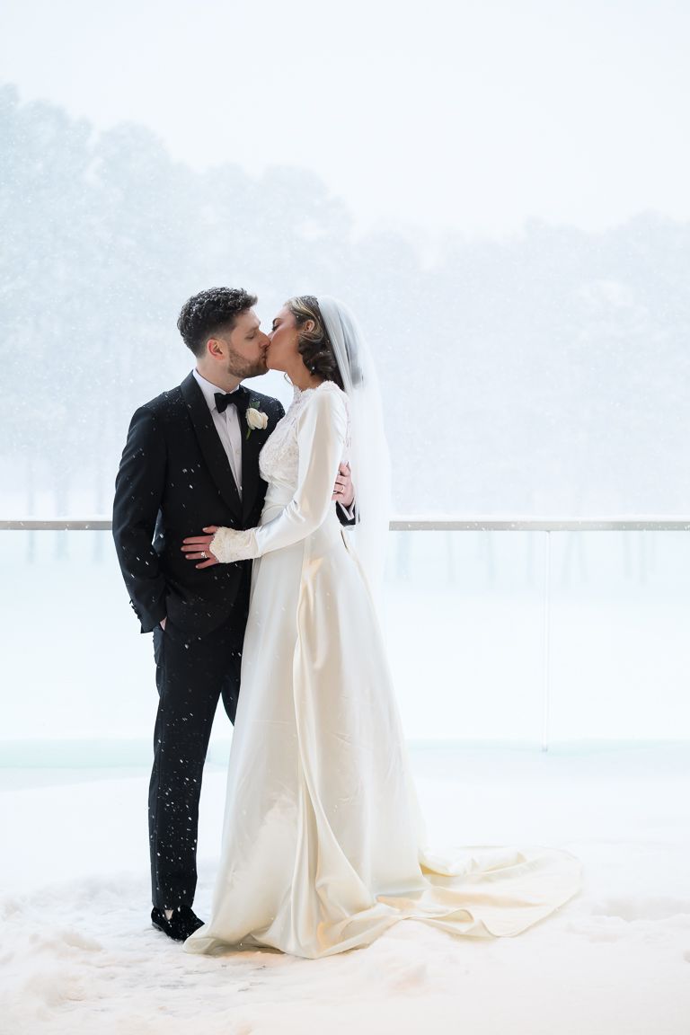 A romantic winter wedding photo of a couple kissing in the snow while wearing formal wedding attire.