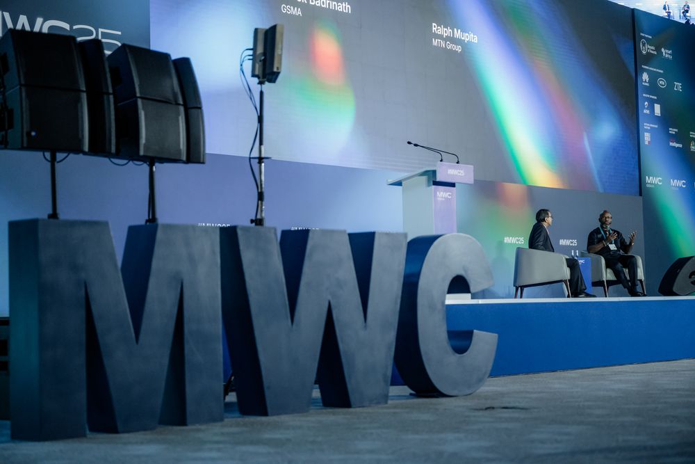Conference stage setup with blue lighting and multiple presentation screens showing business content.