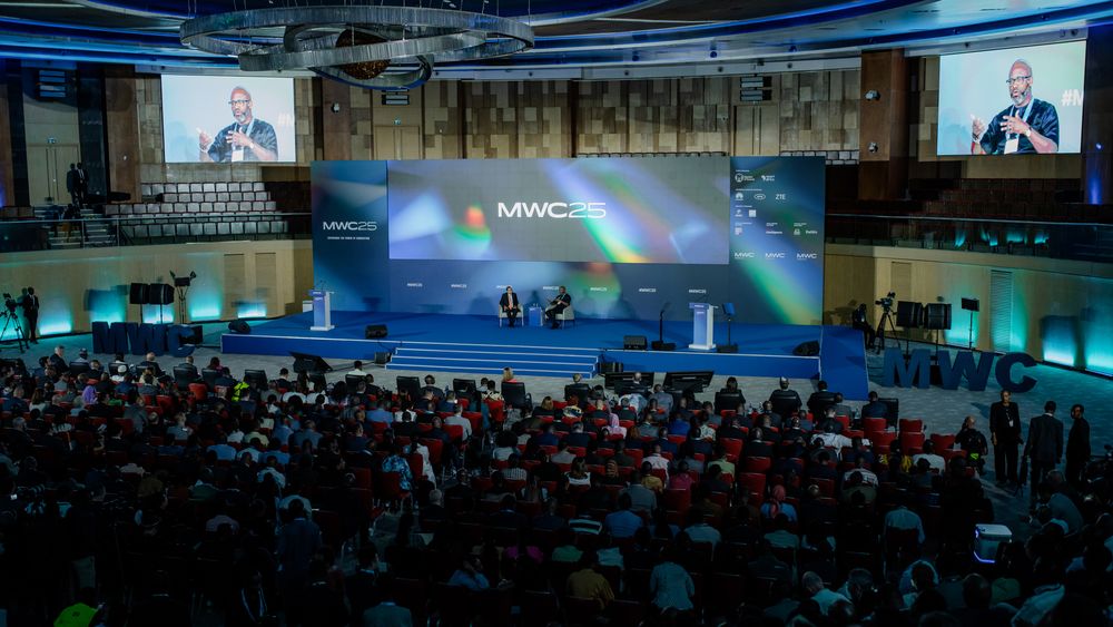Large conference hall with blue lighting and multiple projection screens showing presentations to a packed audience.