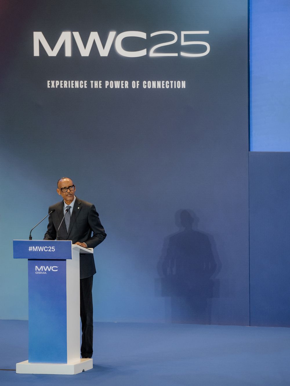 Speaker presenting at MWC25 mobile technology conference from behind a blue podium on stage.