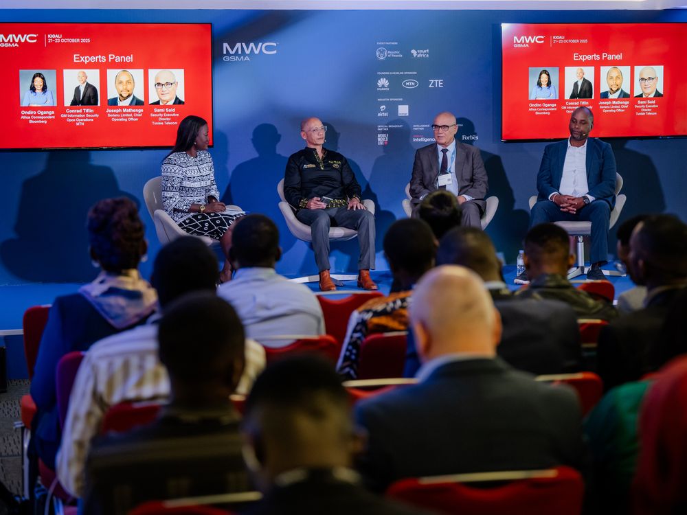 Conference attendees seated in a dimly lit auditorium with red banners and digital displays at a technology event.