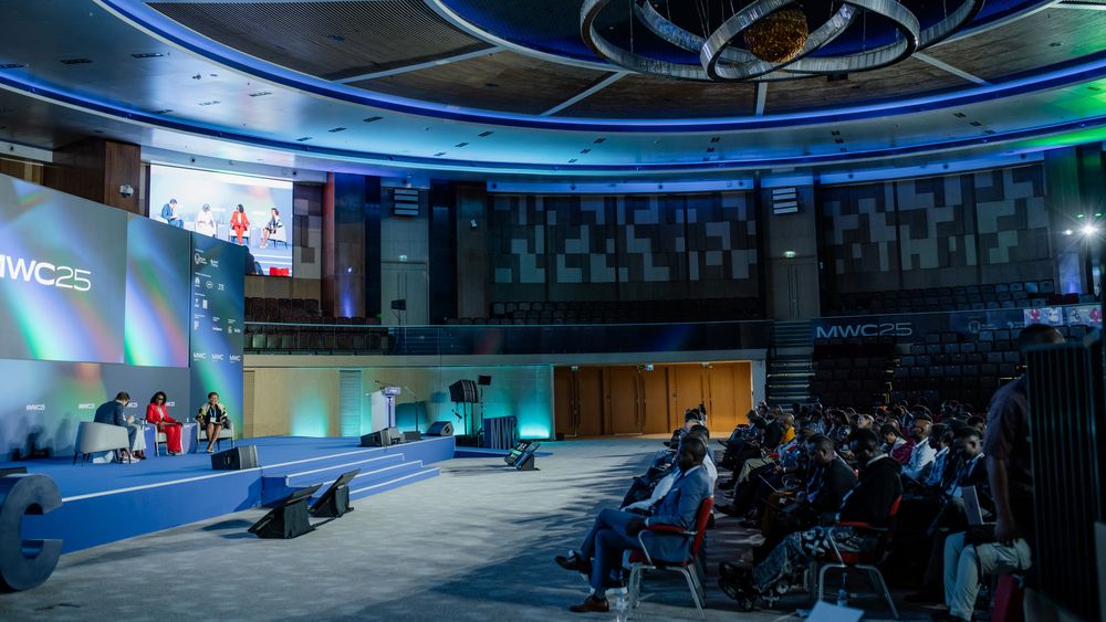 Large conference venue with blue lighting and multiple widescreen displays mounted along curved walls.