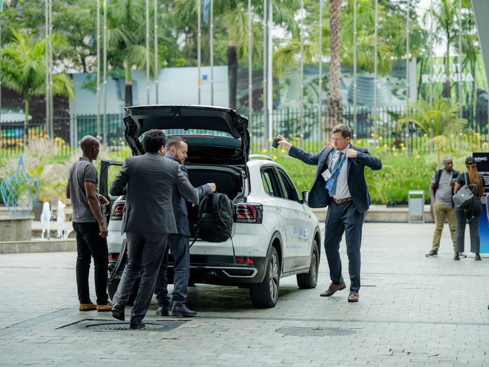 A group of businesspeople gather around an open car trunk in a parking area with greenery in the background.