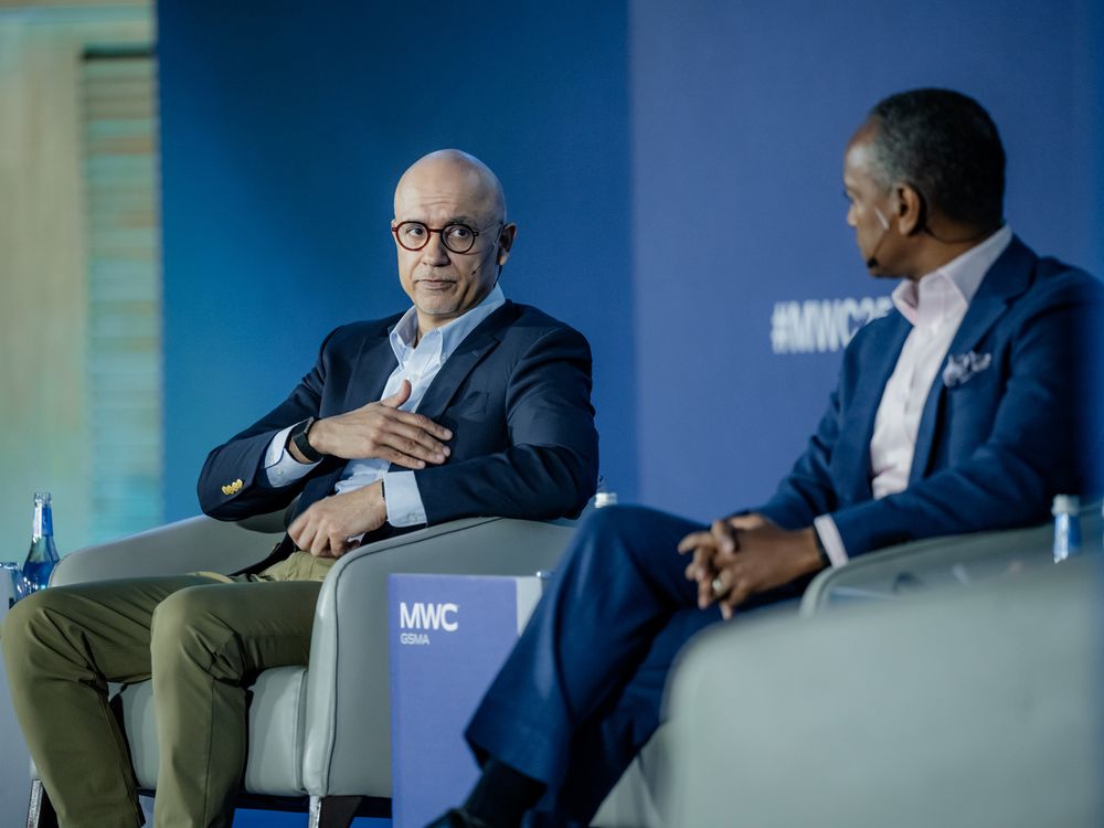Business executives seated in chairs on stage during a professional panel discussion in a blue-lit venue.