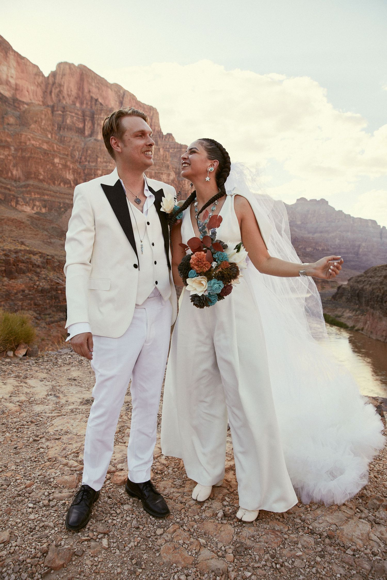 Couple in white attire sharing joyful moments along canyon riverbank.