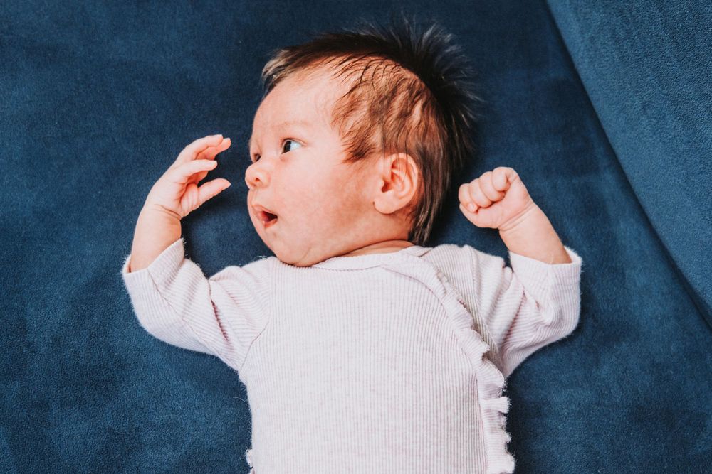 Baby in white ribbed outfit lying on blue blanket with arms raised.