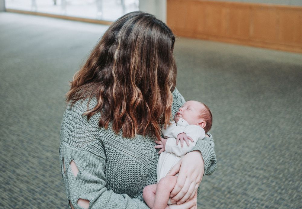 A series of tender moments shows a mother holding a sleeping newborn baby while sitting on grey carpeted floor.