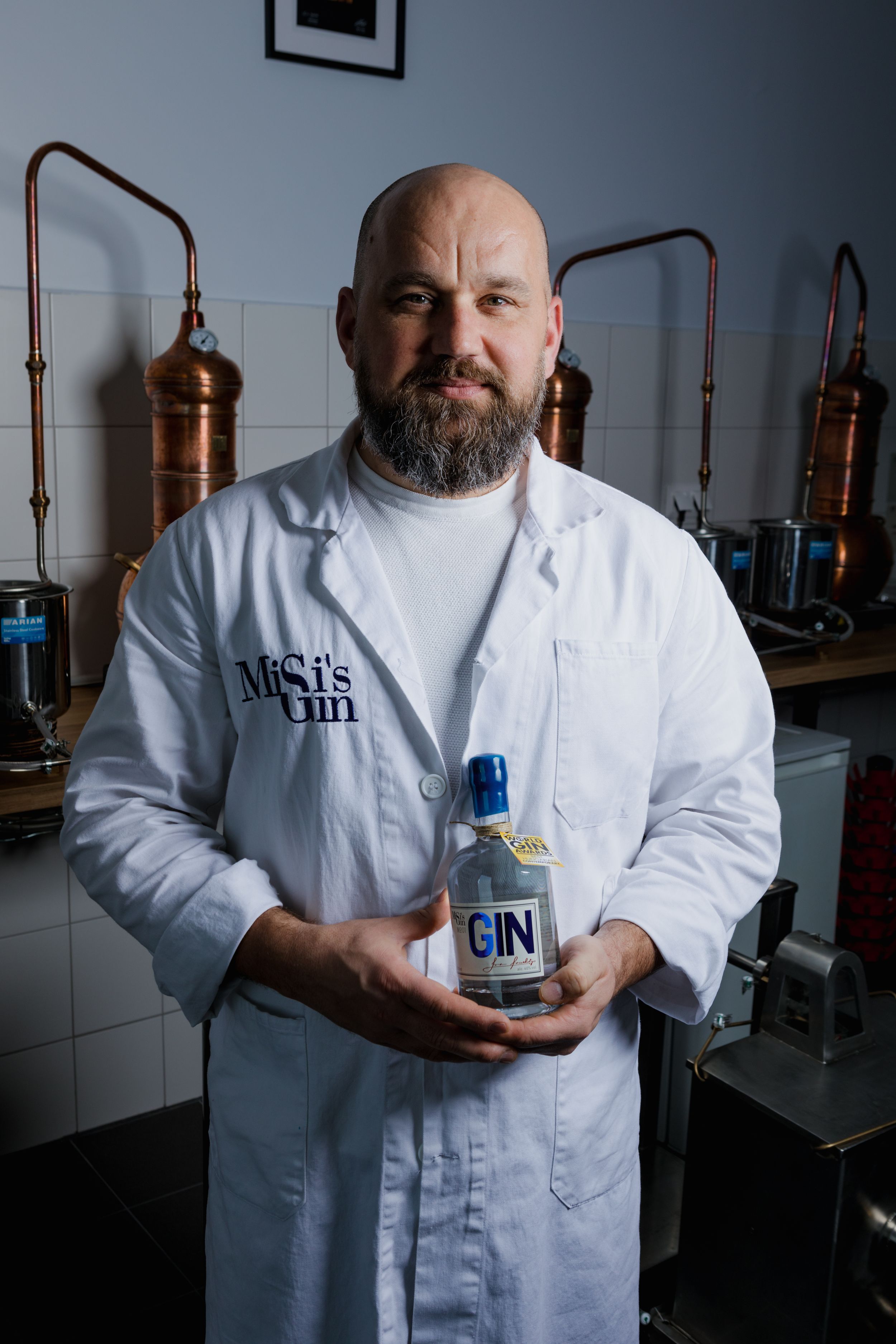 A distillery worker in white coat holding a bottle of gin with copper stills in the background.