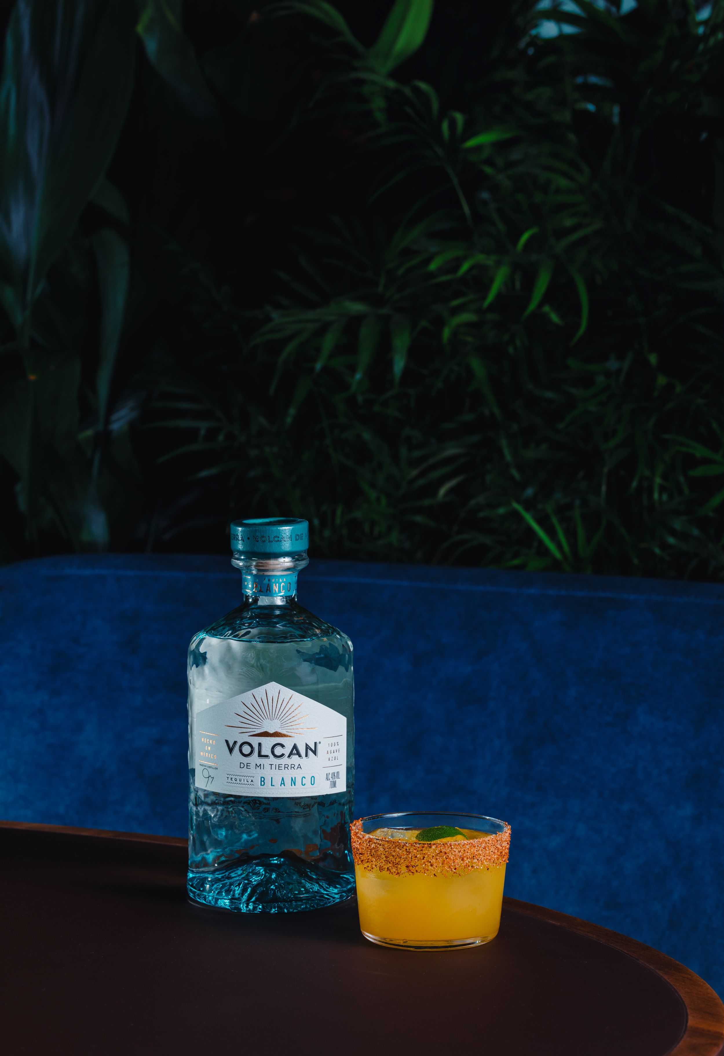 A bottle of Volcan tequila next to an orange cocktail against a blue background.