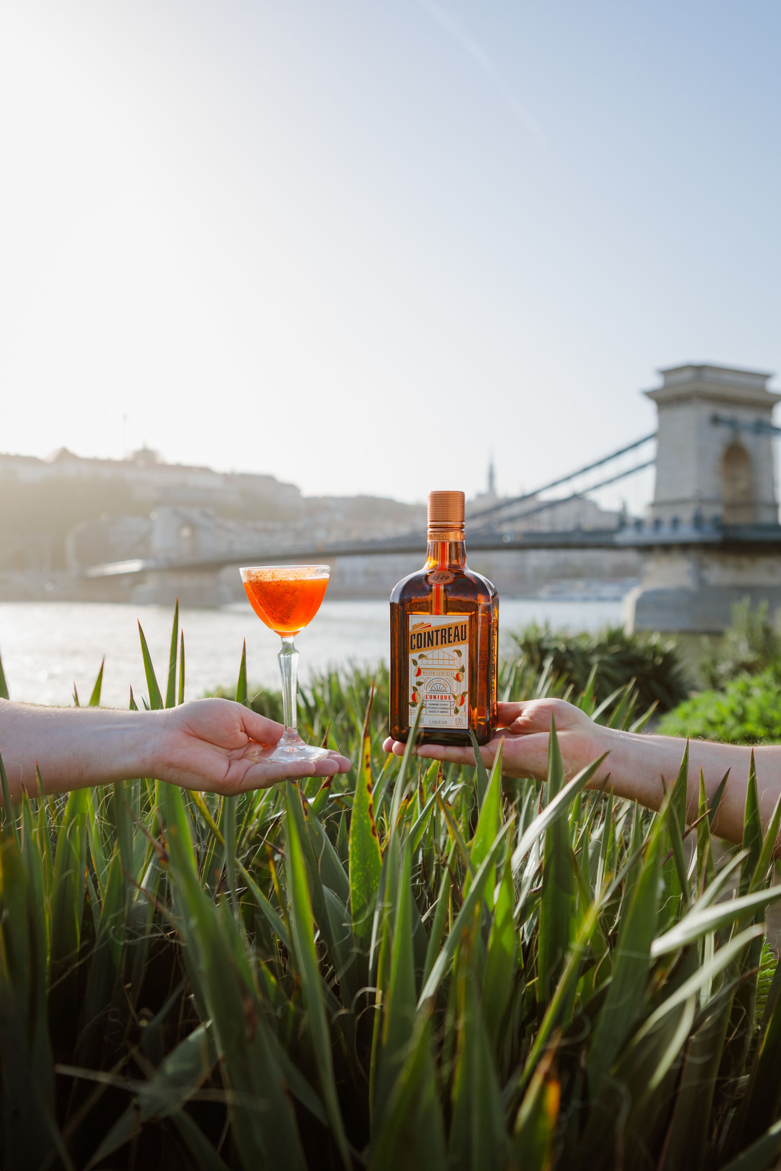 A bottle and cocktail glass being held against the backdrop of the Chain Bridge in Budapest at sunset.