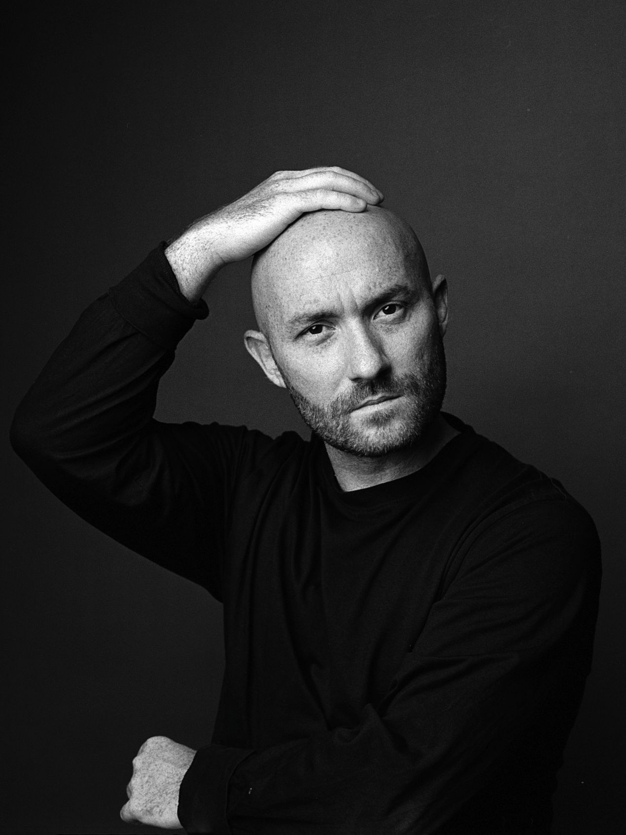 Black and white portrait of a bald man with beard in black sweater, hand on head, studio background.