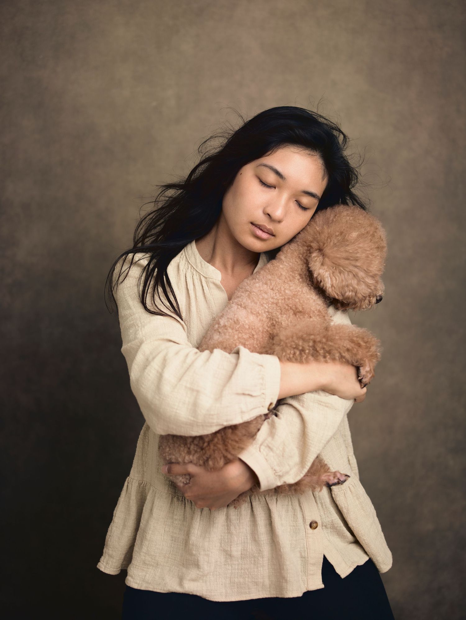 Young woman with dark hair gently hugging a fluffy brown poodle against a warm studio backdrop.