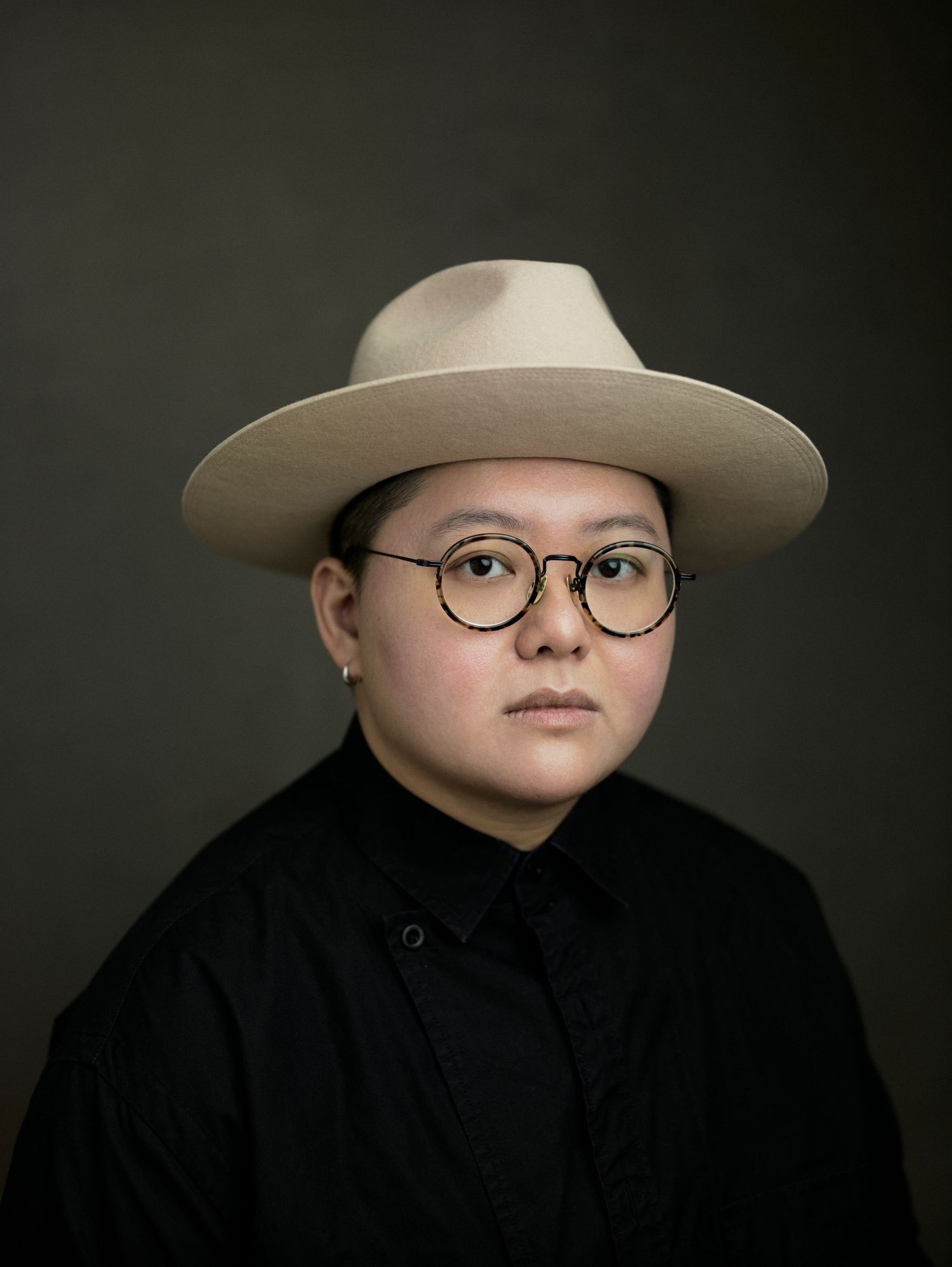 Portrait of a person wearing round glasses and a wide-brim beige hat, dressed in black, against a dark background.