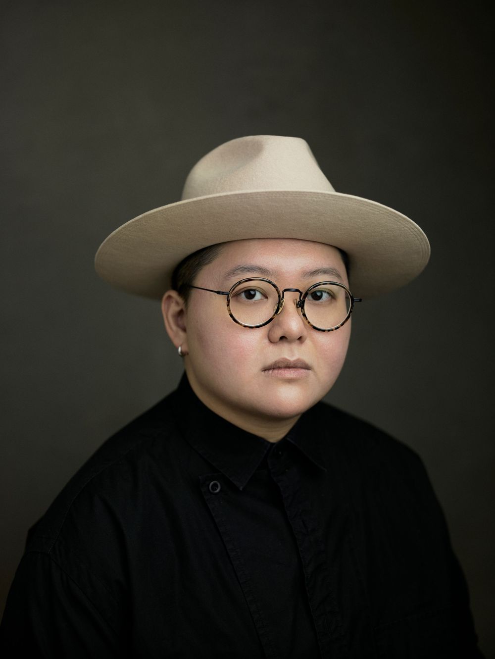 Portrait of a person wearing round glasses and a wide-brim beige hat, dressed in black, against a dark background.