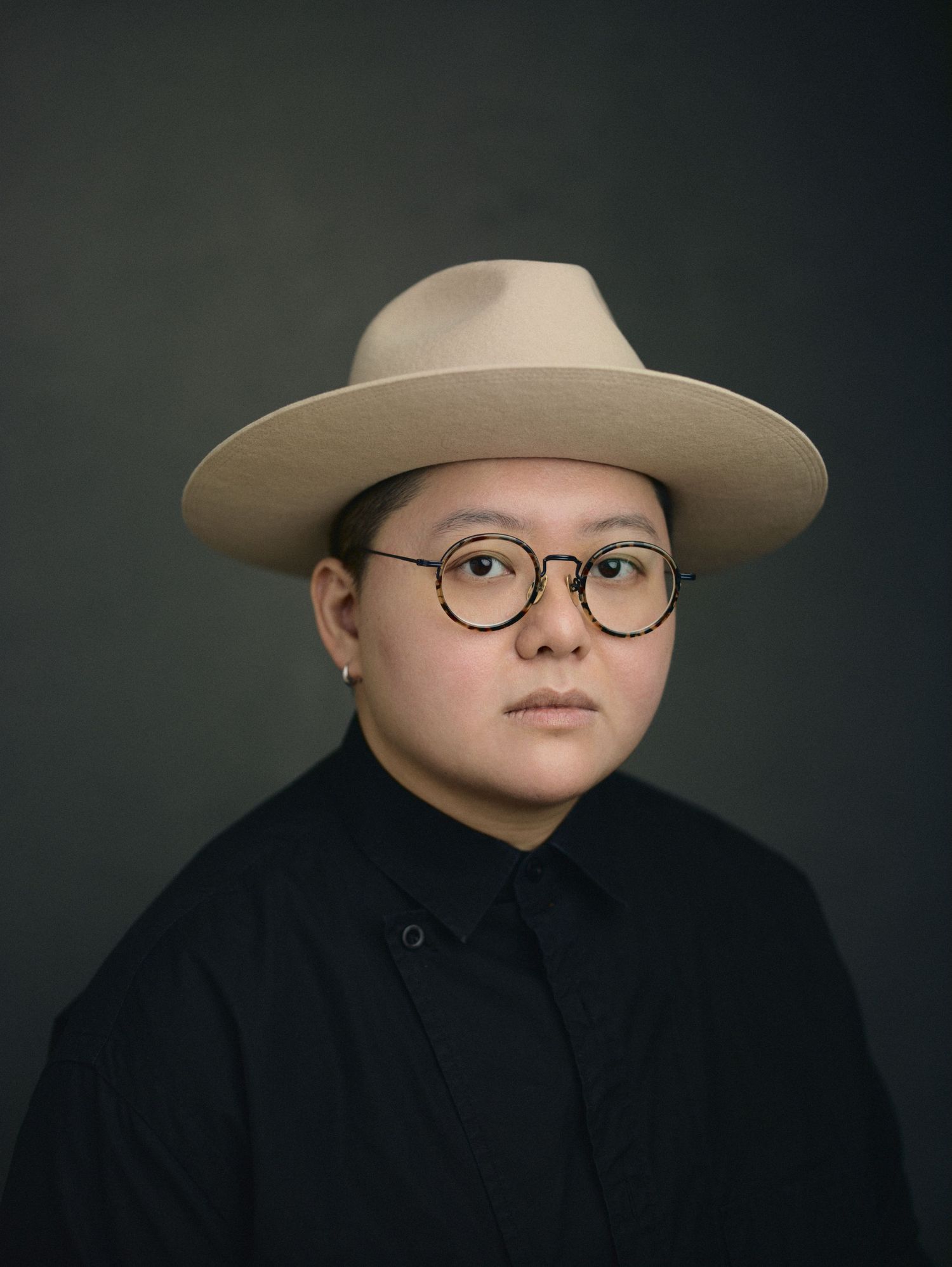 Person wearing round glasses and a wide-brim beige hat, dressed in black, posed against a dark studio background.