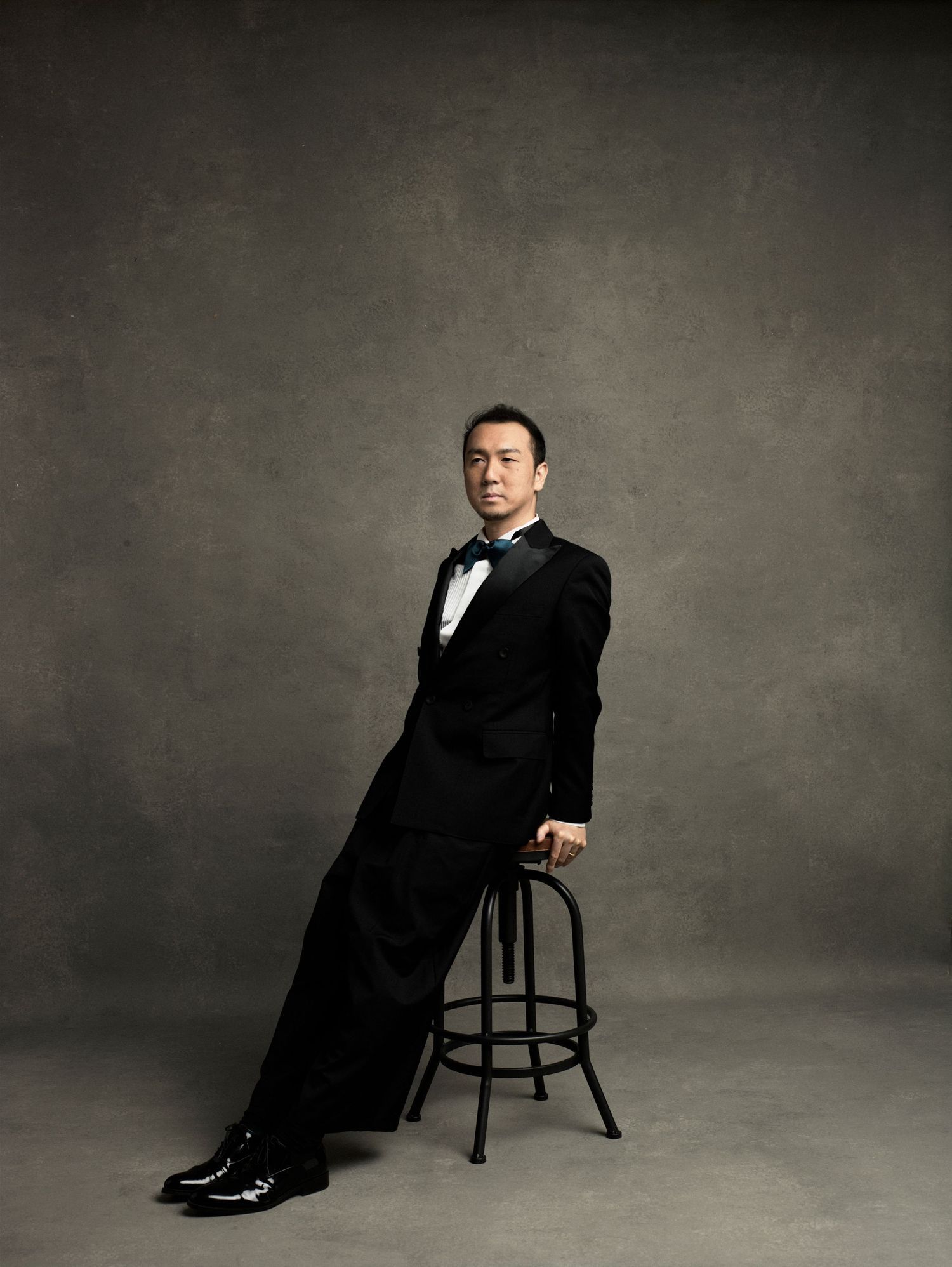 Man in black tuxedo and bow tie poses elegantly on a bar stool against a gray studio background.