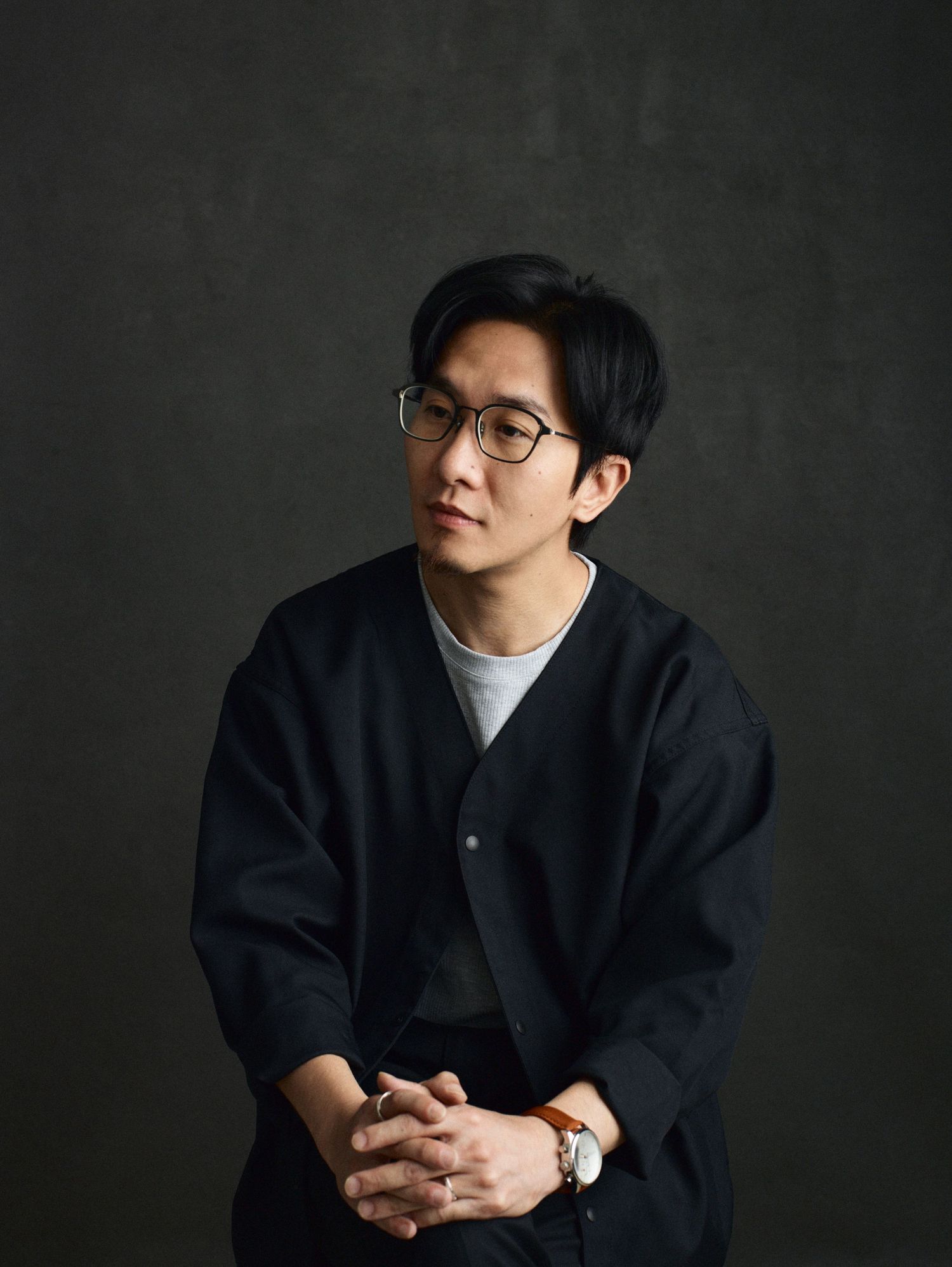 Young Asian man with glasses wearing a black cardigan, posing thoughtfully with hands clasped against a dark background.