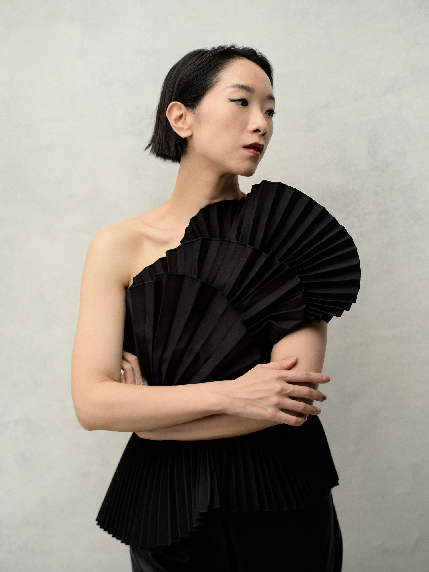 Woman in black pleated avant-garde one-shoulder top poses gracefully against neutral gray background.