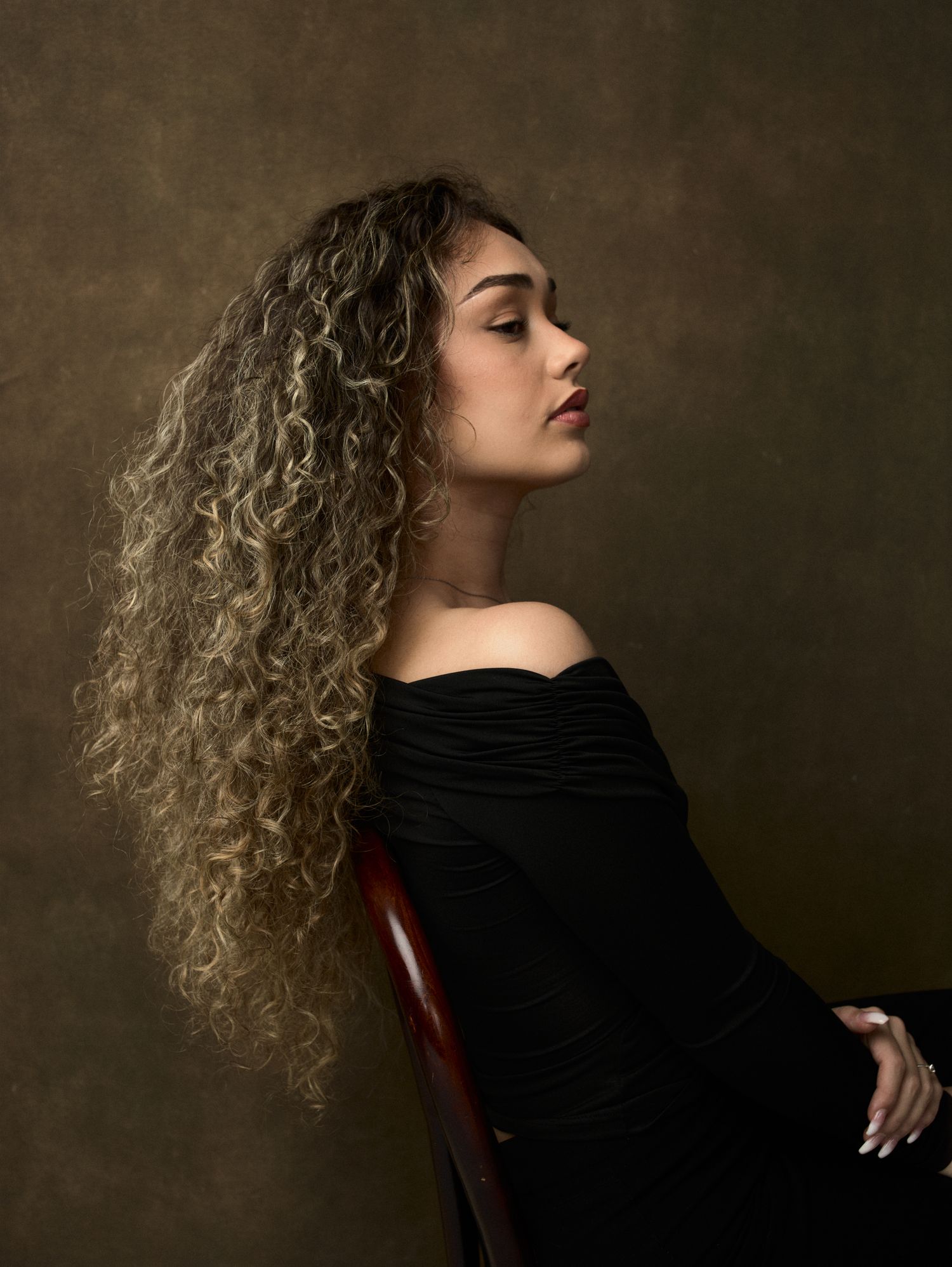 Young woman with long curly blonde-highlighted hair poses in black off-shoulder top against dark background.