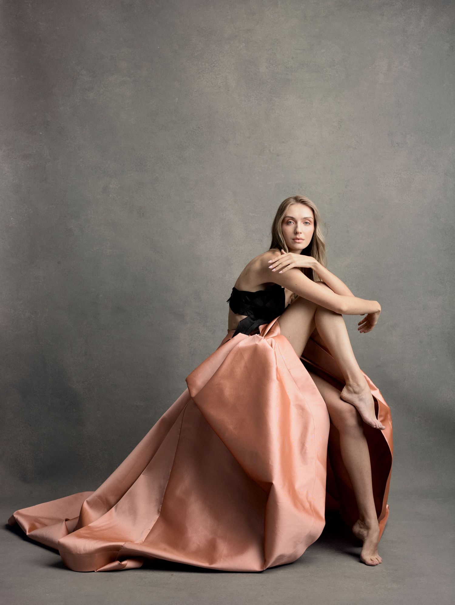 A woman in a black strapless top and flowing peach satin skirt poses gracefully seated on a gray studio backdrop.
