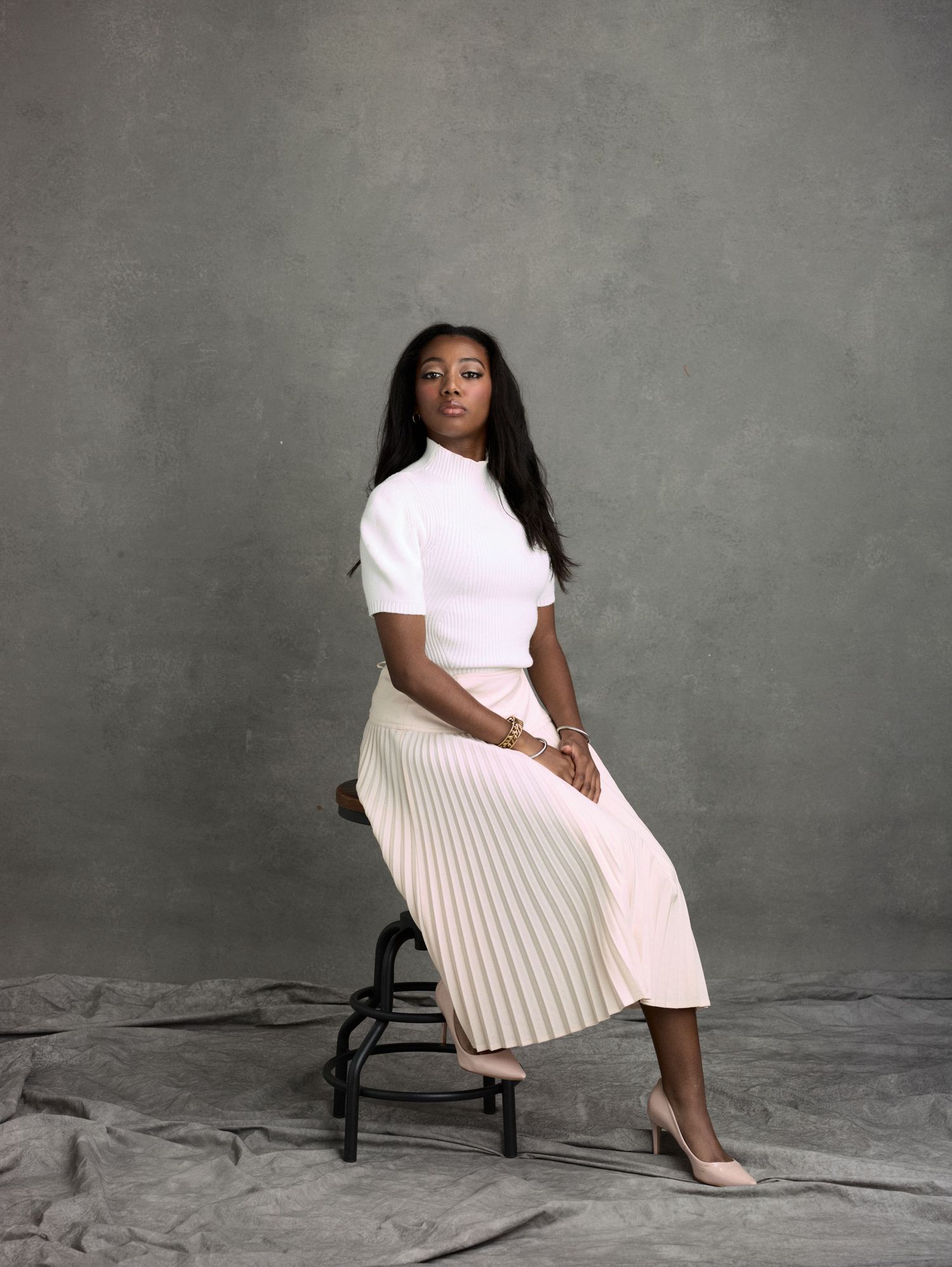 Woman in white top and pleated cream midi skirt seated on stool against gray backdrop in studio portrait.