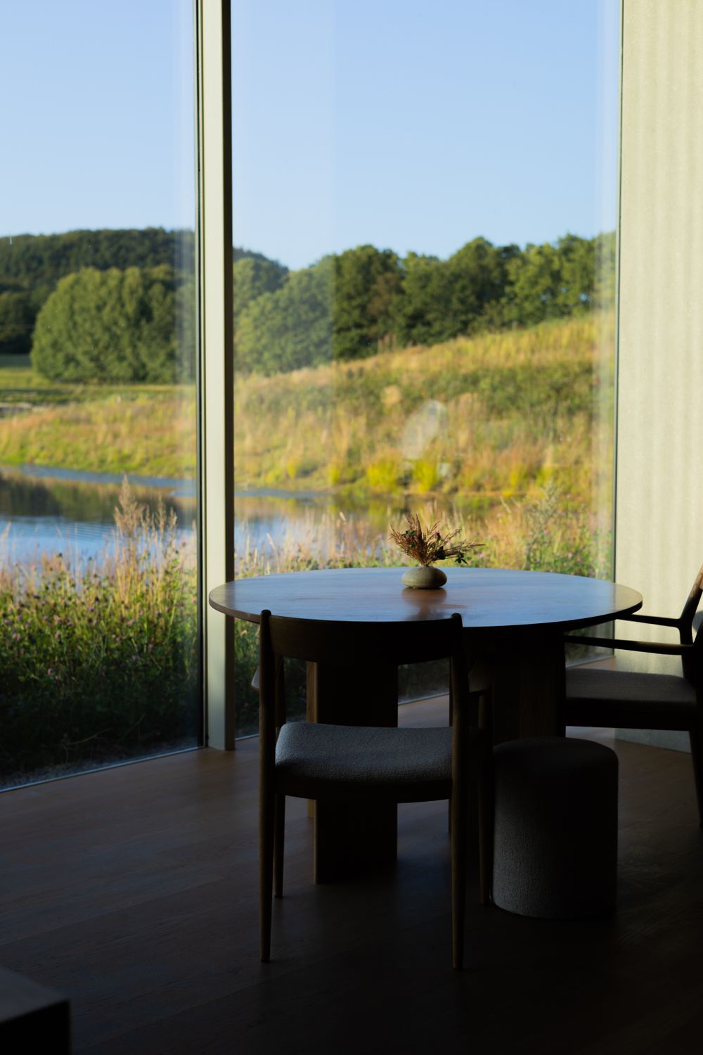 Round wooden table by large window overlooking scenic lake and forest landscape.