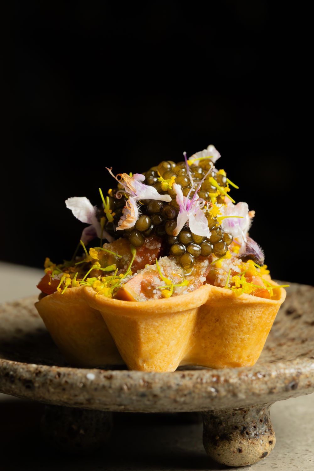 A small pastry tart filled with delicate garnishes and edible flowers on a textured stone plate.
