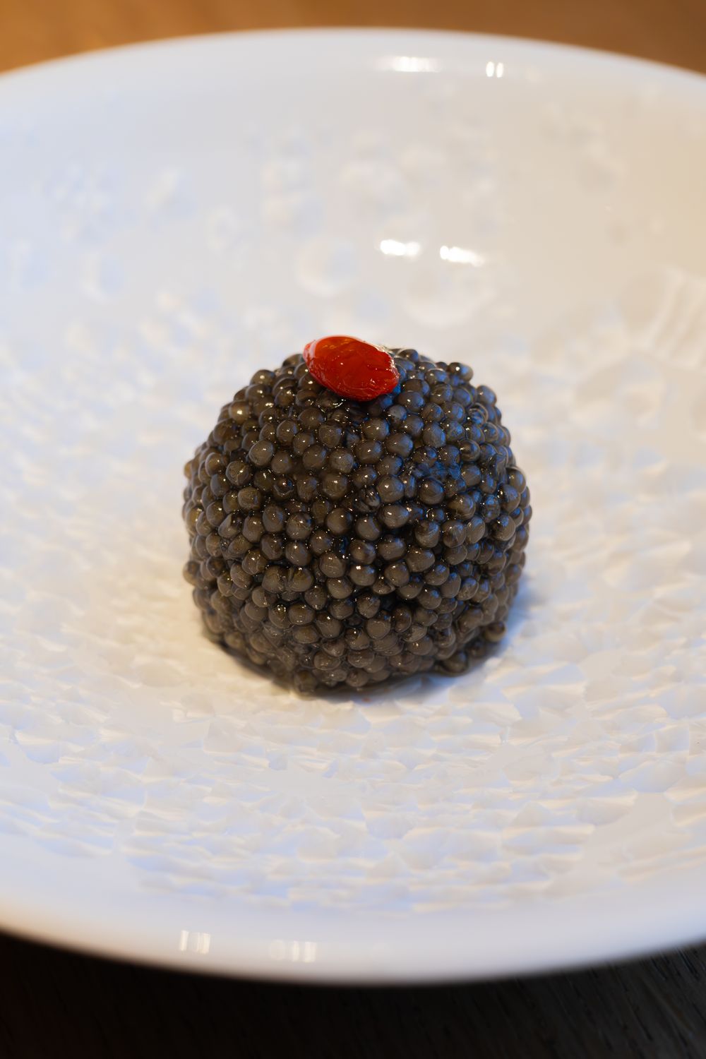 Black caviar ball garnished with a small red topping served on a textured white plate.