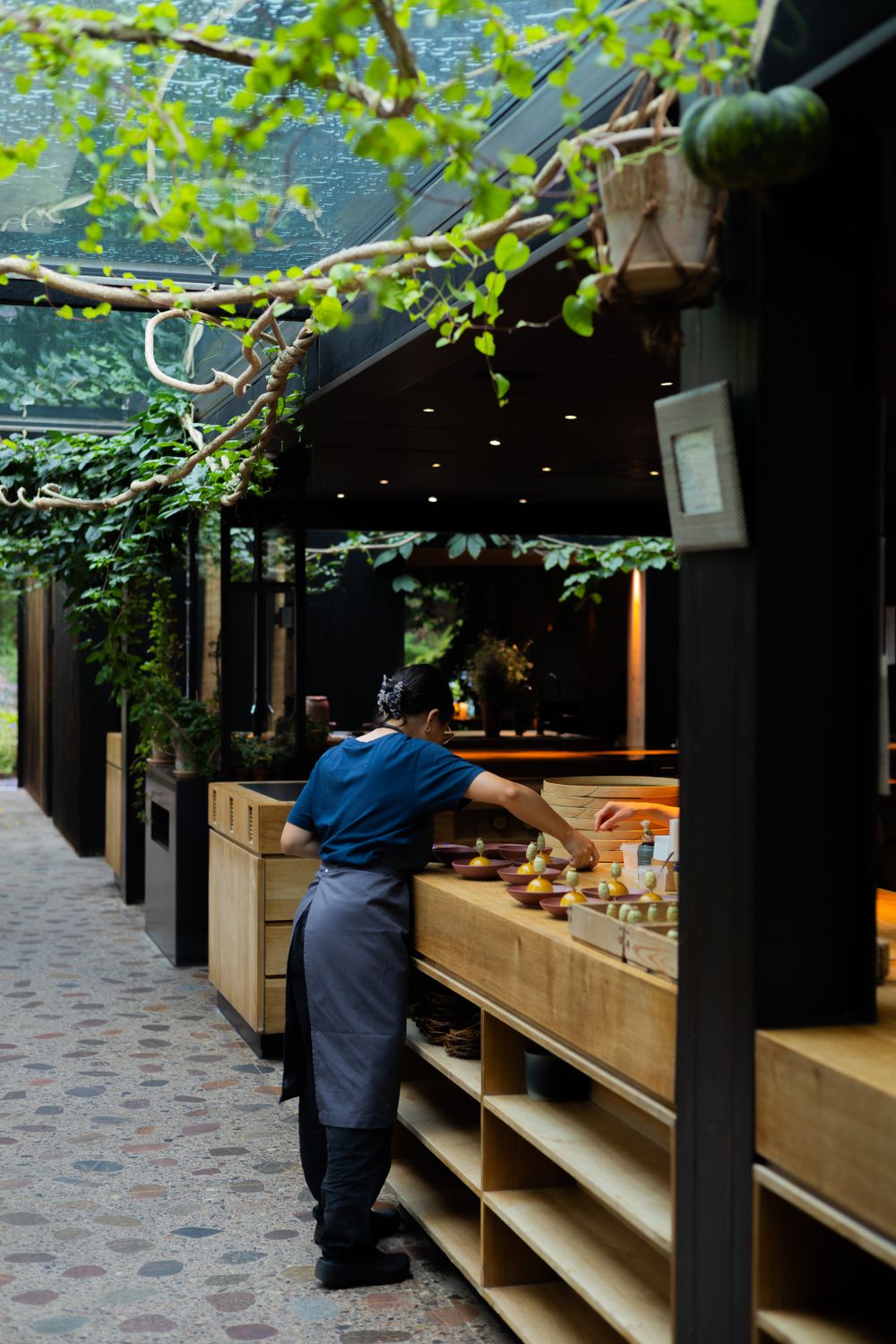 Staff member arranging food items at a wooden counter under a covered outdoor dining area with hanging vines.