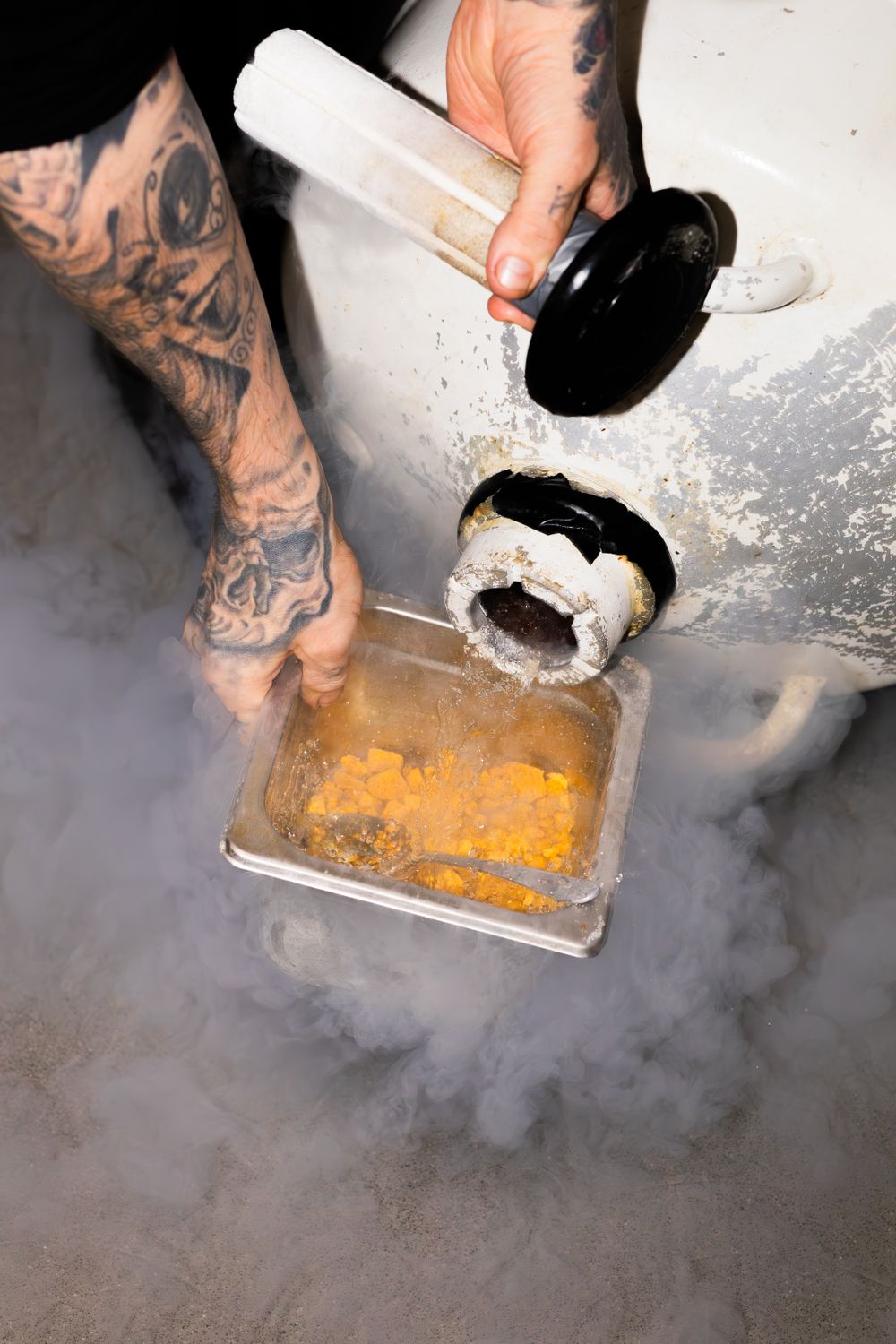 Tattooed hands pouring ingredients into a container surrounded by theatrical smoke.