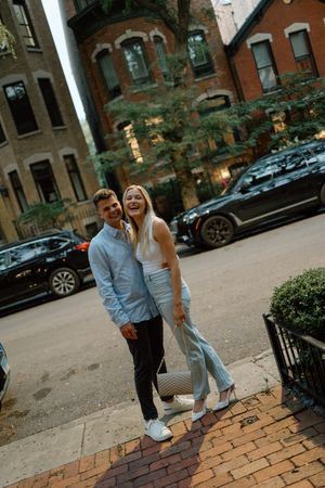 Couple in casual attire pose together on a charming brick-lined street with brownstone buildings.