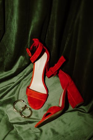 Red high heel sandals and wedding rings arranged on green velvet fabric.