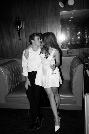 A romantic black and white photo shows a couple in elegant attire sharing an intimate moment on a leather booth in a dimly lit venue.