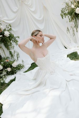A person in a flowing white wedding gown twirls gracefully outdoors among willow trees and floral decorations.