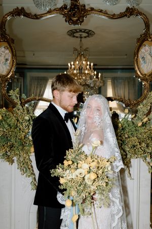 An elegant indoor wedding ceremony with ornate gold mirror, chandelier and abundant floral decorations in yellow and green tones.