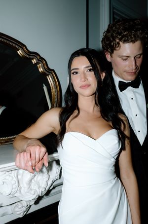 A couple in formal attire poses together at an elegant event, with the woman wearing a white dress.