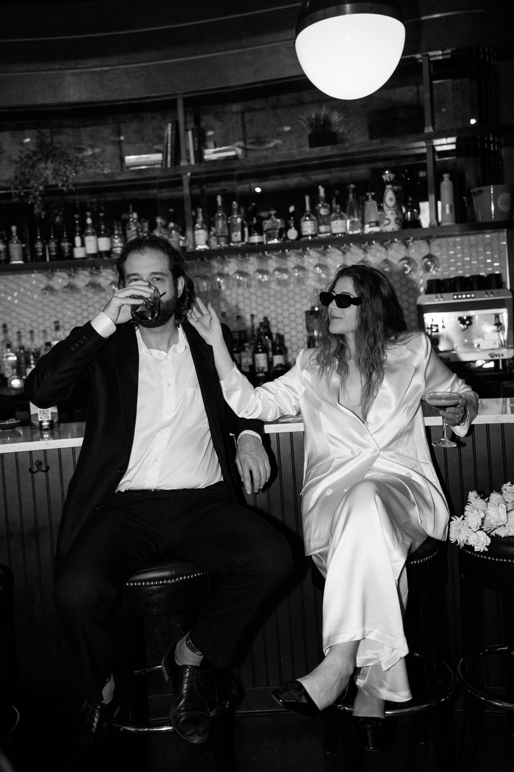 A black and white photo of two people sitting at a bar counter with cocktail bottles in the background.