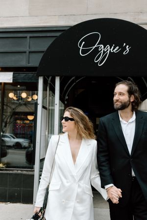 A couple in stylish outfits holding hands while walking past Ozzie's restaurant with black awning overhead.