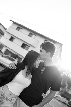 A romantic black and white photo of a young couple embracing outdoors in front of a building at sunset.