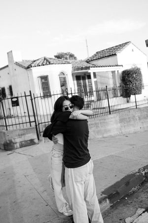 A tender black and white photograph of a couple sharing a warm embrace on a sidewalk outside a house with iron fence.