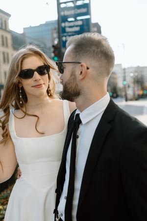 Stylish couple in formal attire share an intimate moment on a city street with urban buildings in the background.