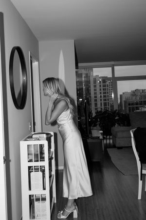 Elegant figure in white dress poses in modern apartment with city views through large windows.