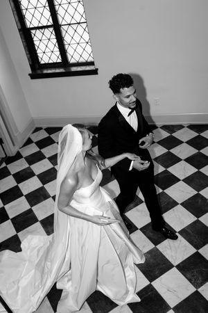 Black and white photo of bride and groom dancing on checkered floor beneath a lattice window.