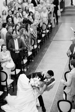 Black and white photo of bride and groom sharing a romantic dip kiss in the aisle surrounded by cheering wedding guests.